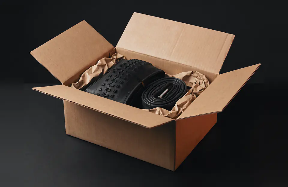 An open cardboard box packed with a folded knobby tire and a rolled inner tube, cushioned with brown paper