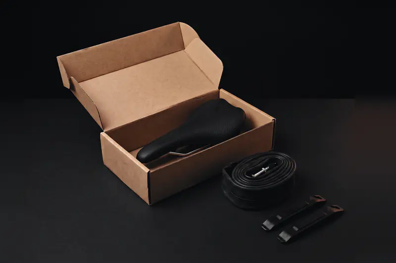 An open shipping box holding a bike saddle, with a rolled inner tube and two tire levers beside it